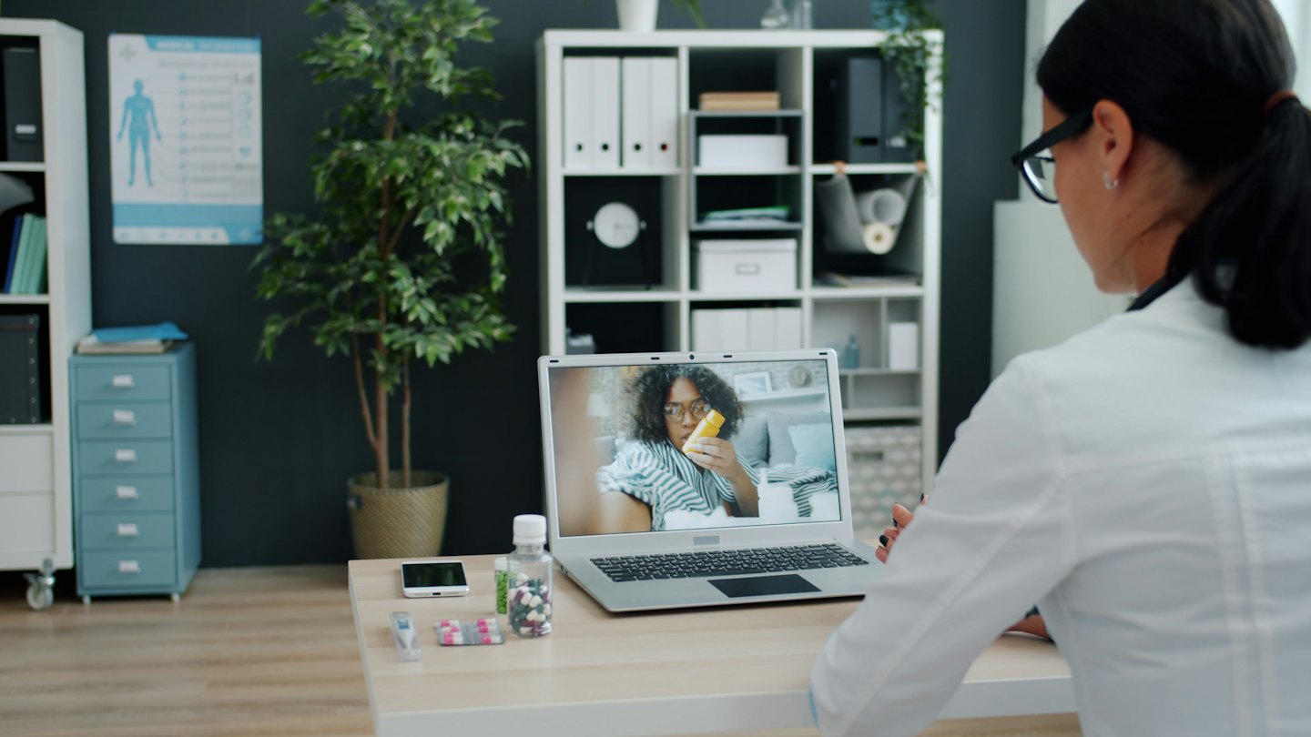 Lékař na telemedicínském hovoru s pacientem, který drží lahvičku se žlutými pilulkami.
